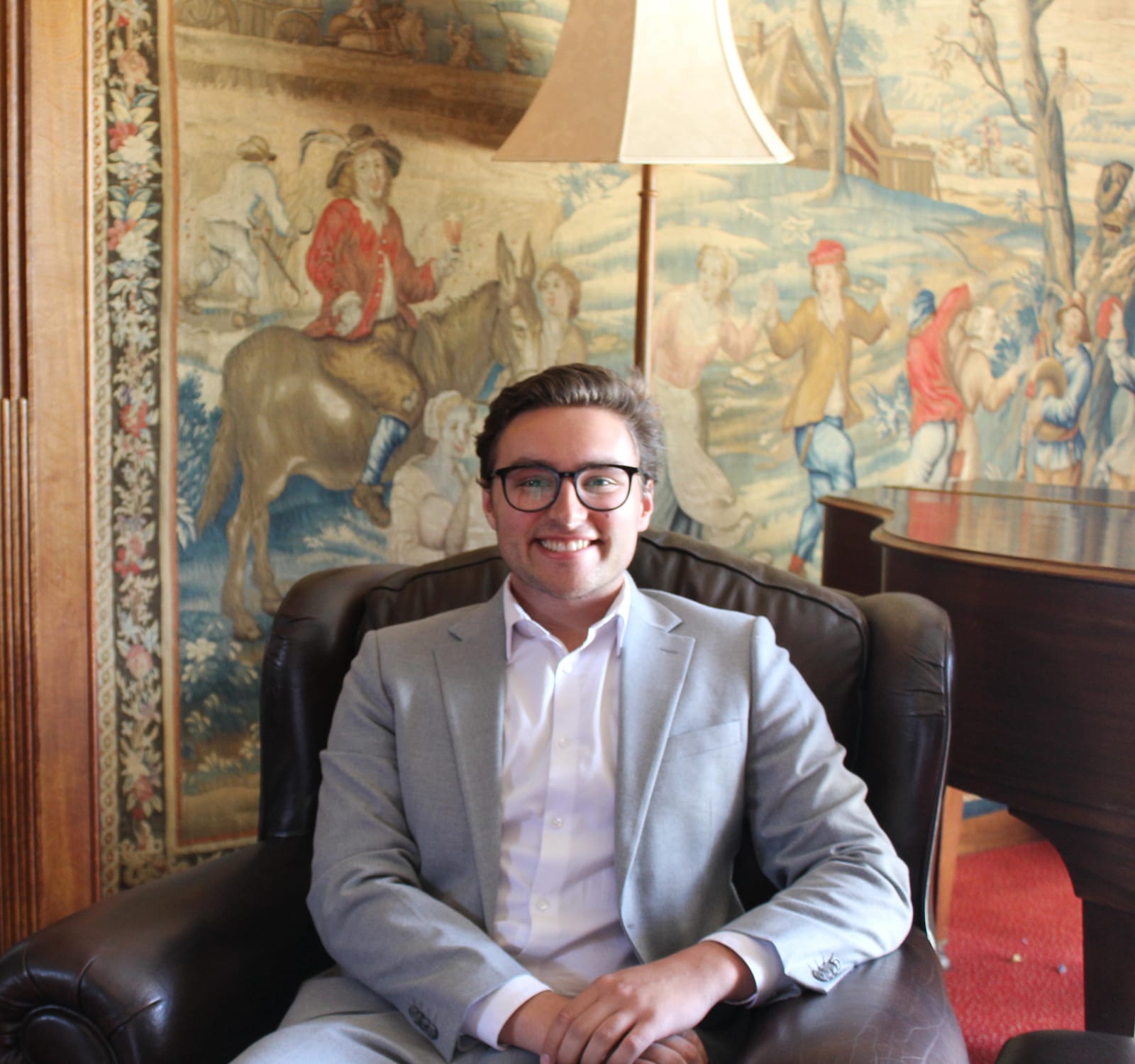 Stephen Weinstock in a light grey blazer, smiling in a leather armchair in front of a large tapestry