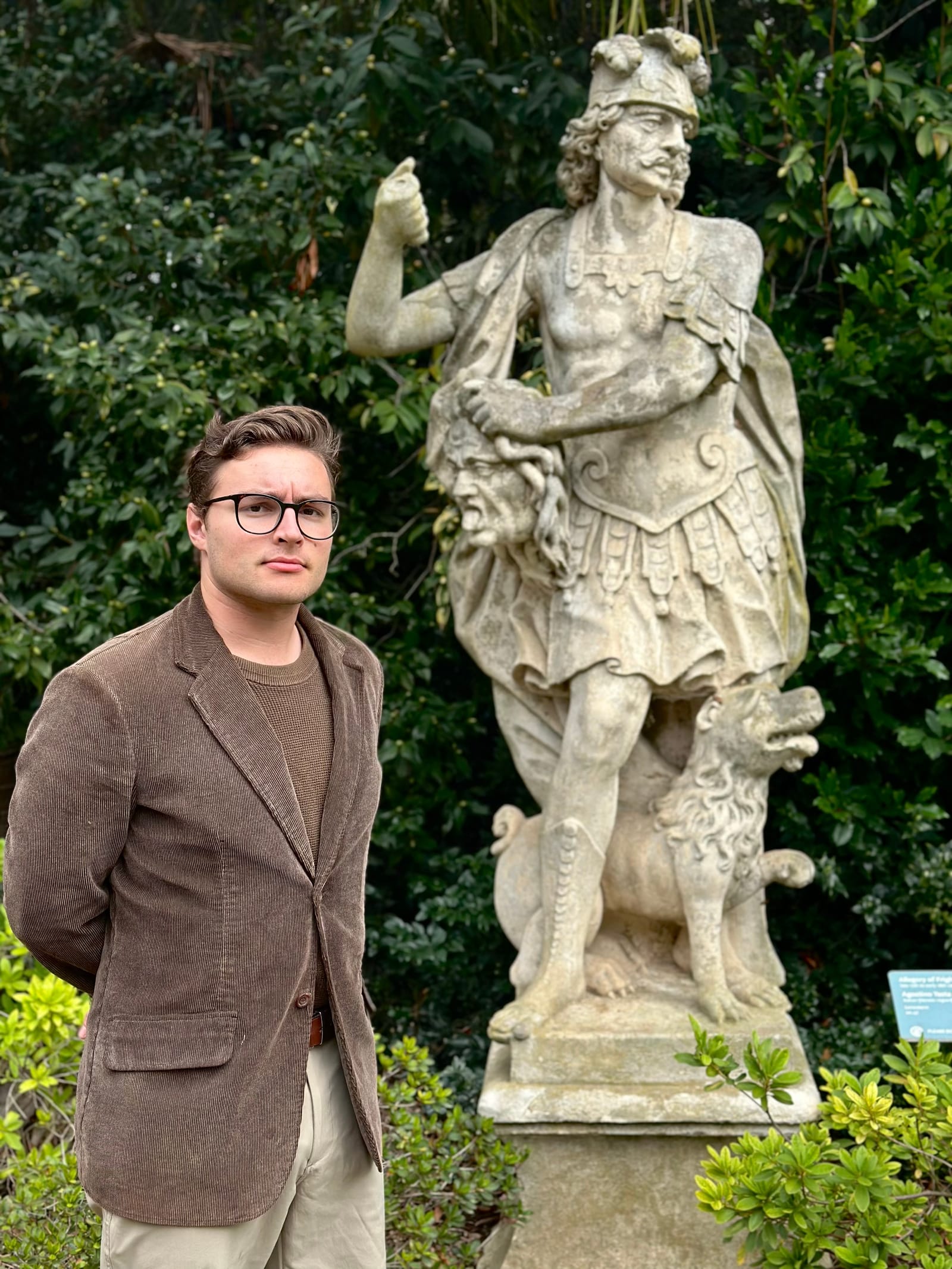 Stephen Weinstock in glasses and a brown corduroy blazer, standing beside a stone statue in a garden