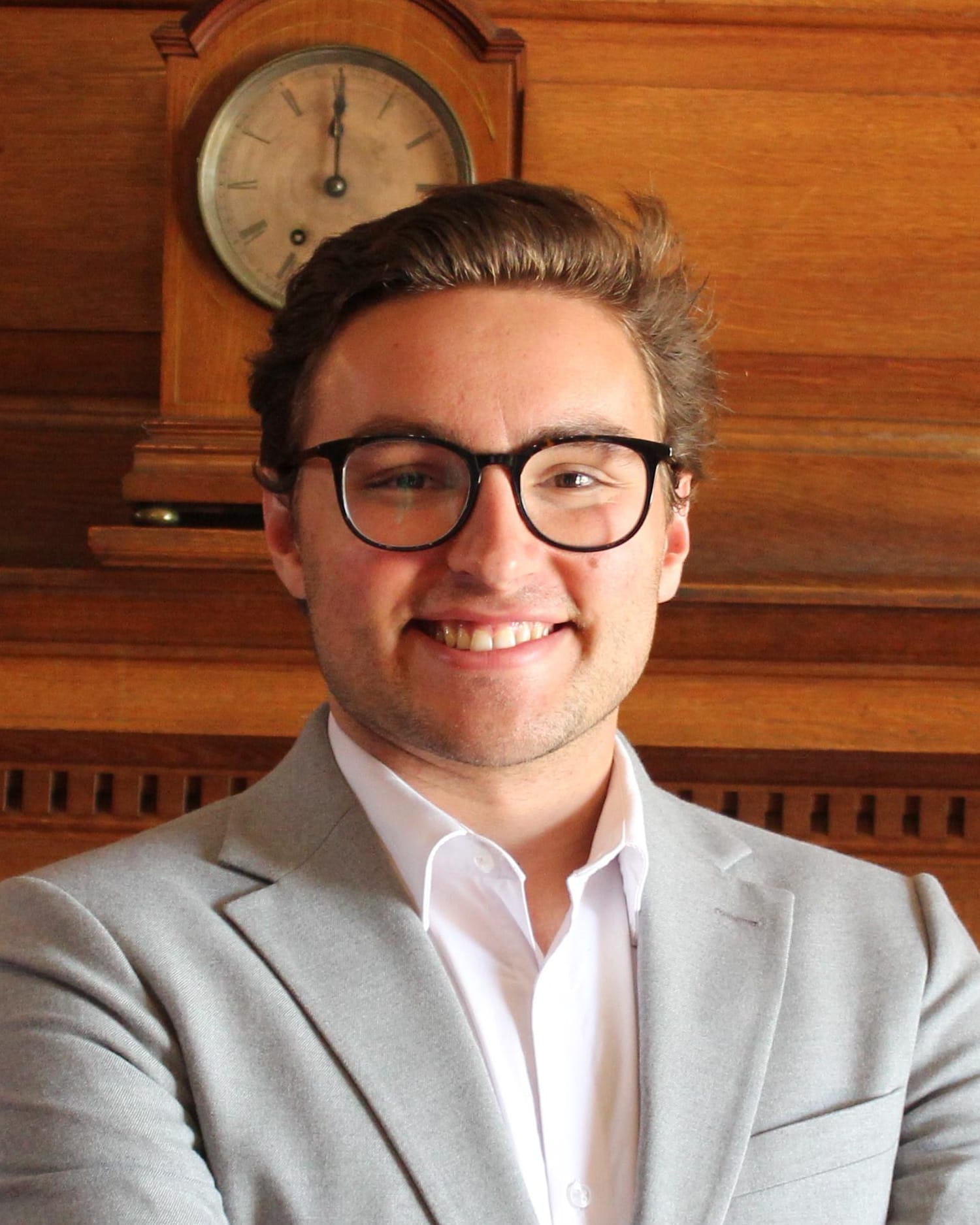 Stephen Weinstock in glasses, a light grey blazer and white shirt, smiling in front of wood panelling and a mantel clock