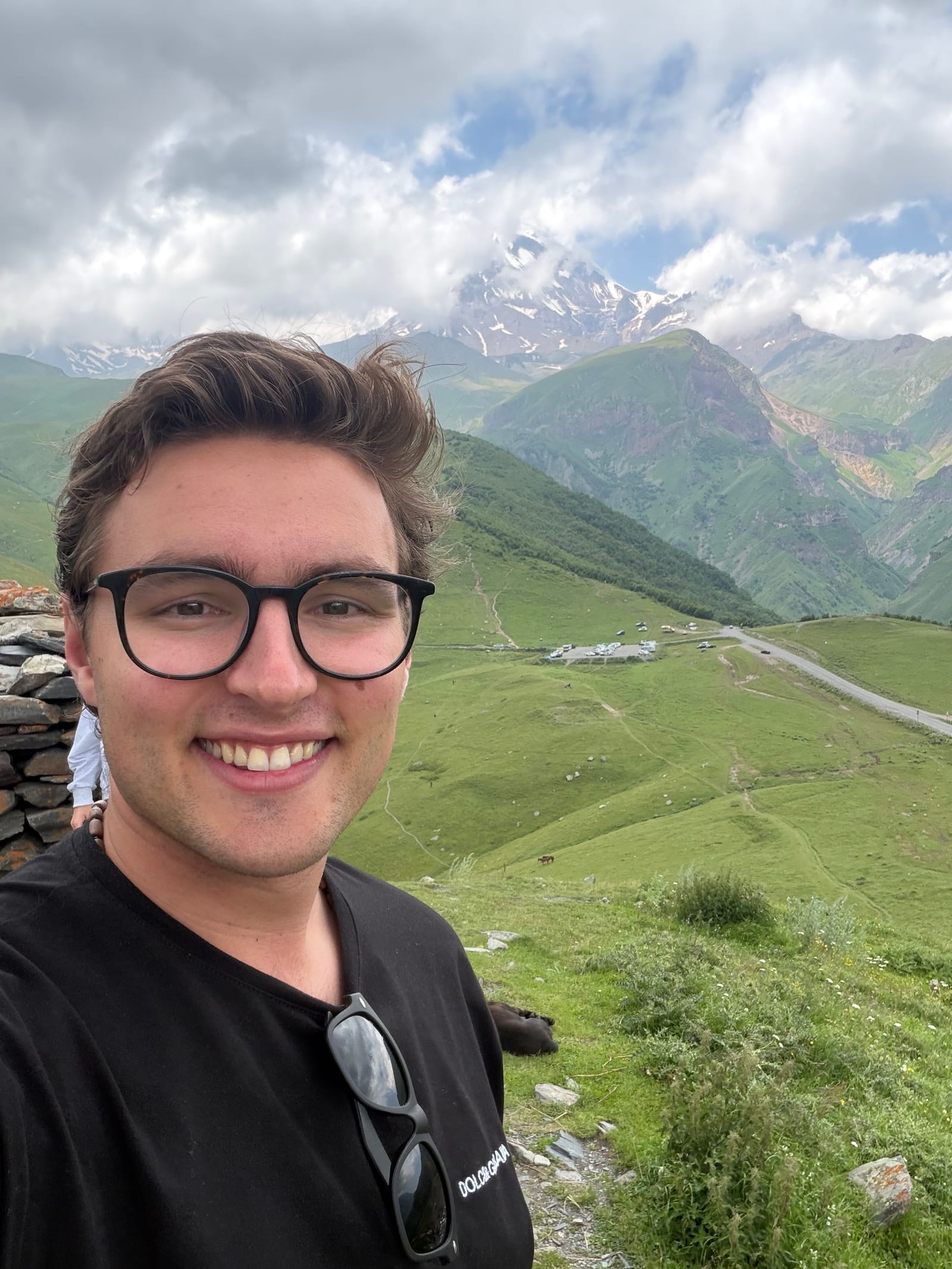 Stephen Weinstock smiling in glasses, with a green valley and snow-capped mountains behind him