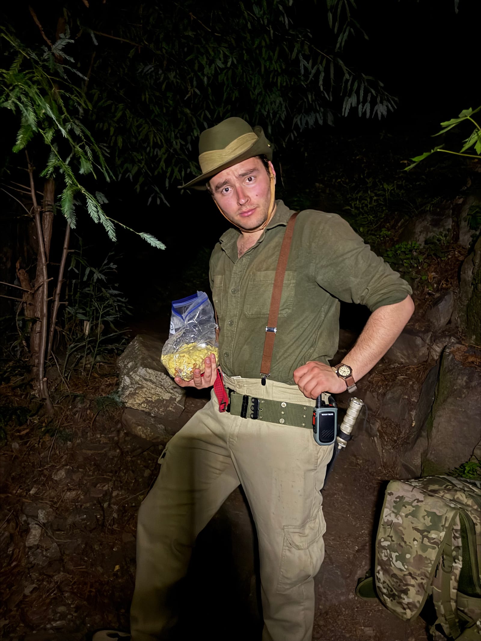 Stephen Weinstock on a night hike in a hat and suspenders, holding a bag of popcorn